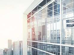 Modern glass office building with reflections of city skyscrapers in the windows, under a bright sky.