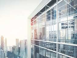 Modern glass office building with reflections of city skyscrapers in the windows, under a bright sky.