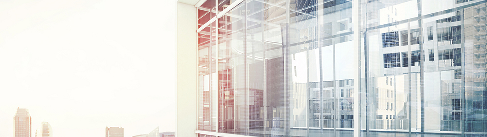 Modern glass office building with reflections of city skyscrapers in the windows, under a bright sky.