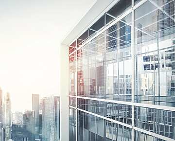 Modern glass office building with reflections of city skyscrapers in the windows, under a bright sky.