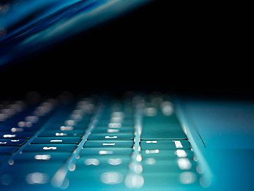 A close-up view of a laptop keyboard illuminated by soft blue light, with the screen partially closed, creating a sleek and futuristic atmosphere.