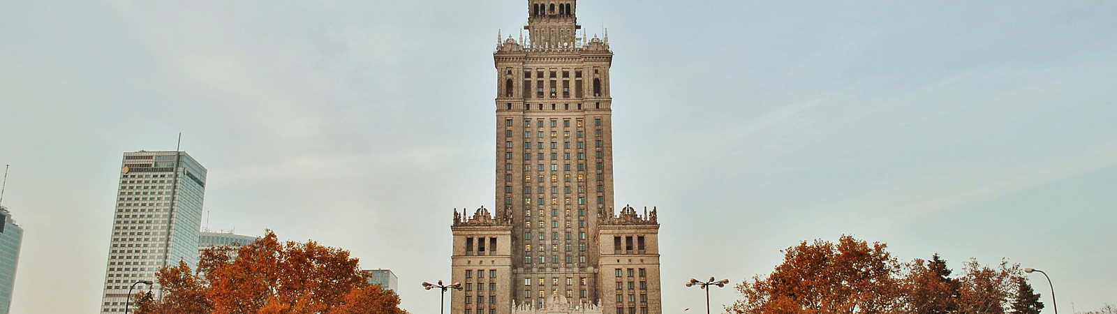 a large building with a clock tower and trees in front of it with Palace of Culture and Science in the background