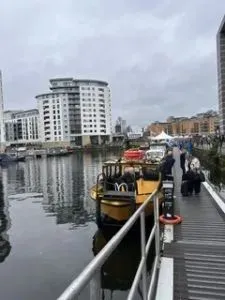 A waterfront walkway with people standing beside the water and buildings in the background.