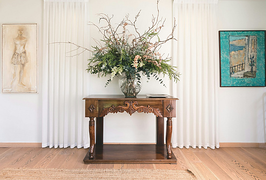 A decorative interior scene featuring a wooden console table with ornate carvings, topped with a large floral arrangement in a glass vase.