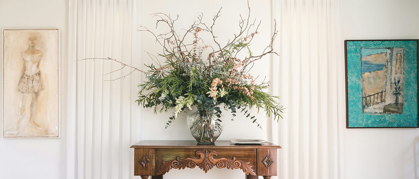 A decorative interior scene featuring a wooden console table with ornate carvings, topped with a large floral arrangement in a glass vase.