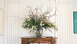 A decorative interior scene featuring a wooden console table with ornate carvings, topped with a large floral arrangement in a glass vase.