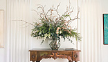 A decorative interior scene featuring a wooden console table with ornate carvings, topped with a large floral arrangement in a glass vase.