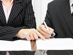 Two people in business attire sitting at a desk, with one person pointing at a document while the other writes on it.