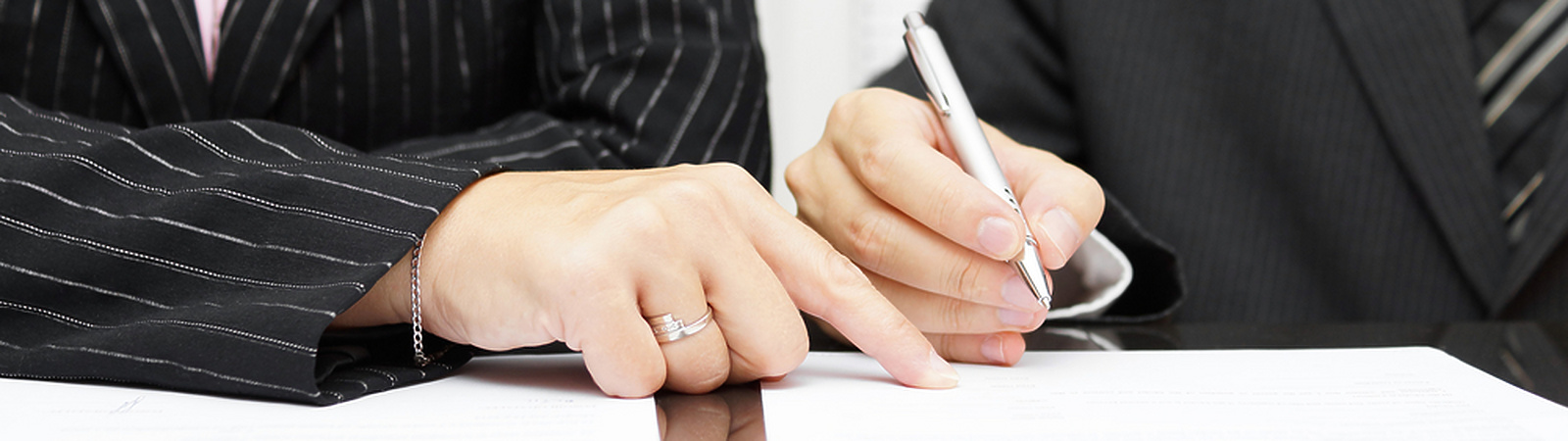 Two people in business attire sitting at a desk, with one person pointing at a document while the other writes on it.