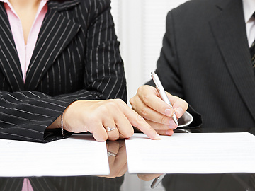 Two people in business attire sitting at a desk, with one person pointing at a document while the other writes on it.