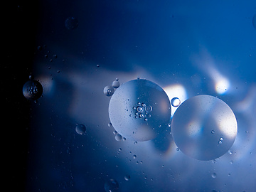 Abstract image of translucent oil droplets suspended in blue liquid, illuminated by light reflections and shadows, creating a soft, ethereal effect.