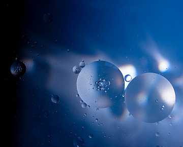 Abstract image of translucent oil droplets suspended in blue liquid, illuminated by light reflections and shadows, creating a soft, ethereal effect.